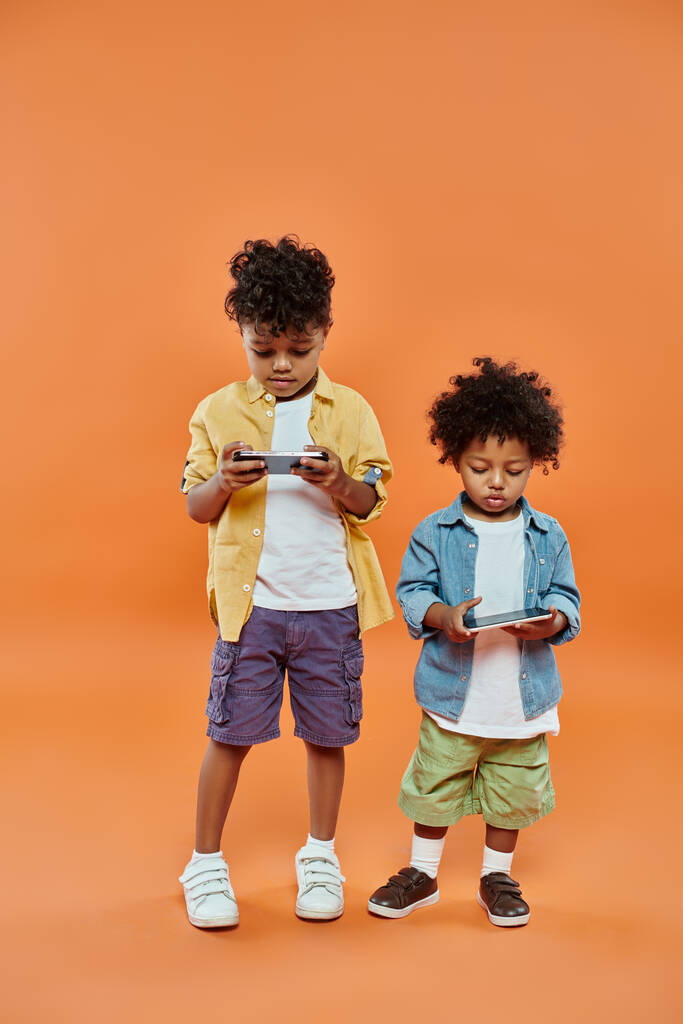 Gülümseyen Afro-Amerikan kardeşler, günlük kıyafetleriyle turuncu üzerinde akıllı telefonlarla oynuyorlar. - Fotoğraf, Görsel