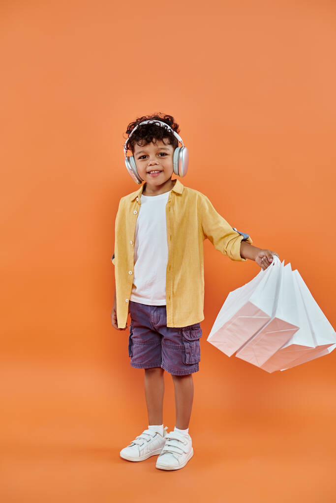 szczęśliwy Afryki amerykański chłopiec słuchając muzyki w słuchawki i trzymając torby na zakupy na pomarańczowy - Zdjęcie, obraz