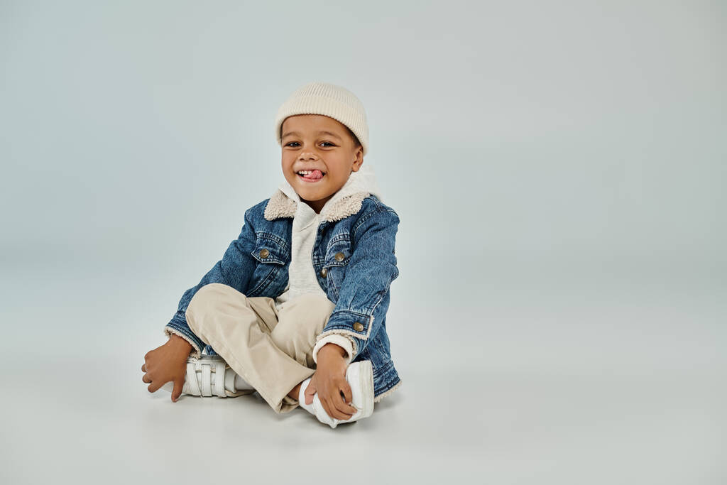 radosny afrykański amerykański przedszkolak chłopiec w stroju zimowym i kapelusz czapka siedzi na szarym tle - Zdjęcie, obraz