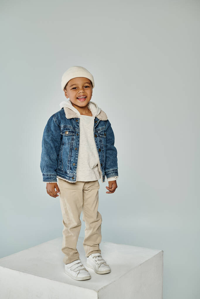 vidám afro-amerikai óvodás fiú téli öltözékben és sapka szürke háttér - Fotó, kép