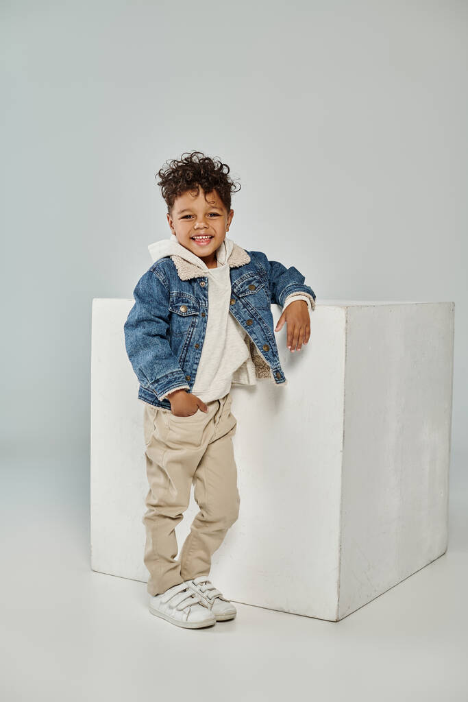 pleased african american boy in winter attire and beanie sitting on concrete cube on grey backdrop - Photo, Image