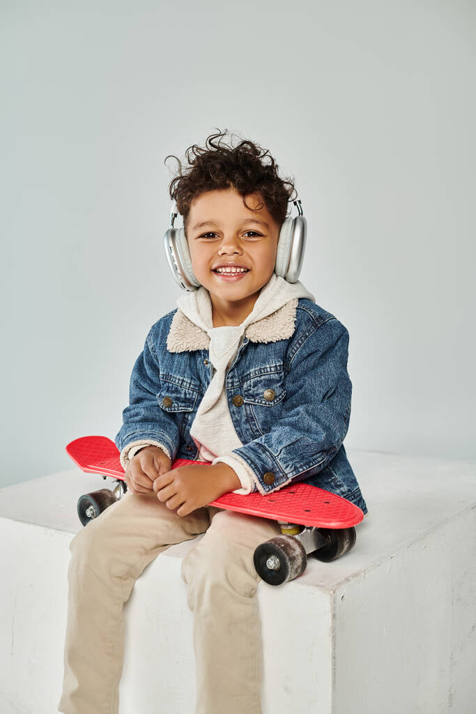happy african american boy in winter attire and headphones sitting on cube with penny board on grey - Photo, Image