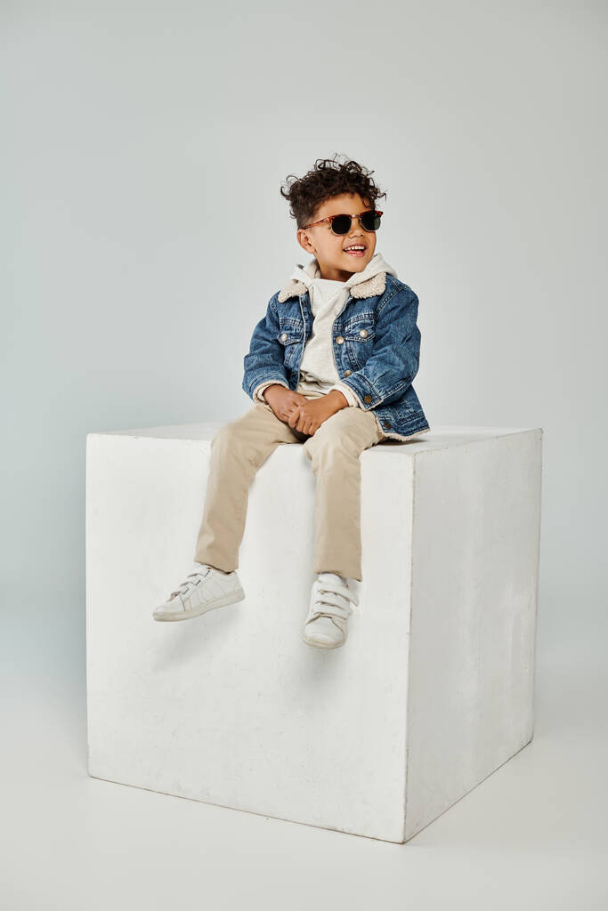 joyful curly african american boy in winter attire and sunglasses sitting on cube on grey backdrop - Photo, Image