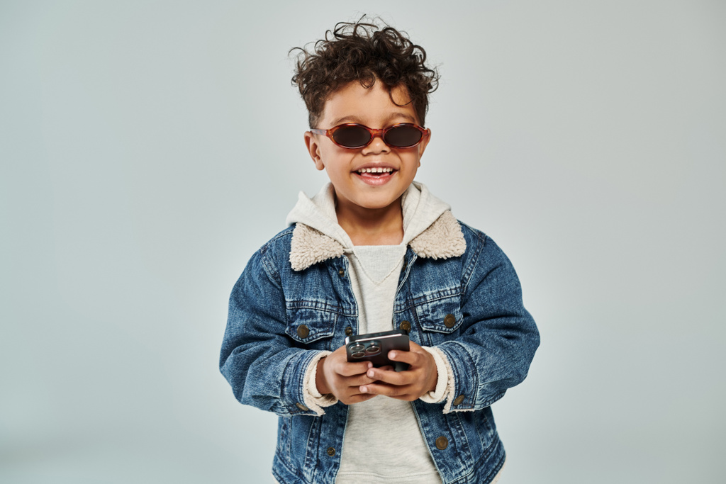 Kışın giyinmiş, güneş gözlüklü mutlu Afrikalı Amerikalı çocuk gri arka planda akıllı telefon kullanıyor. - Fotoğraf, Görsel