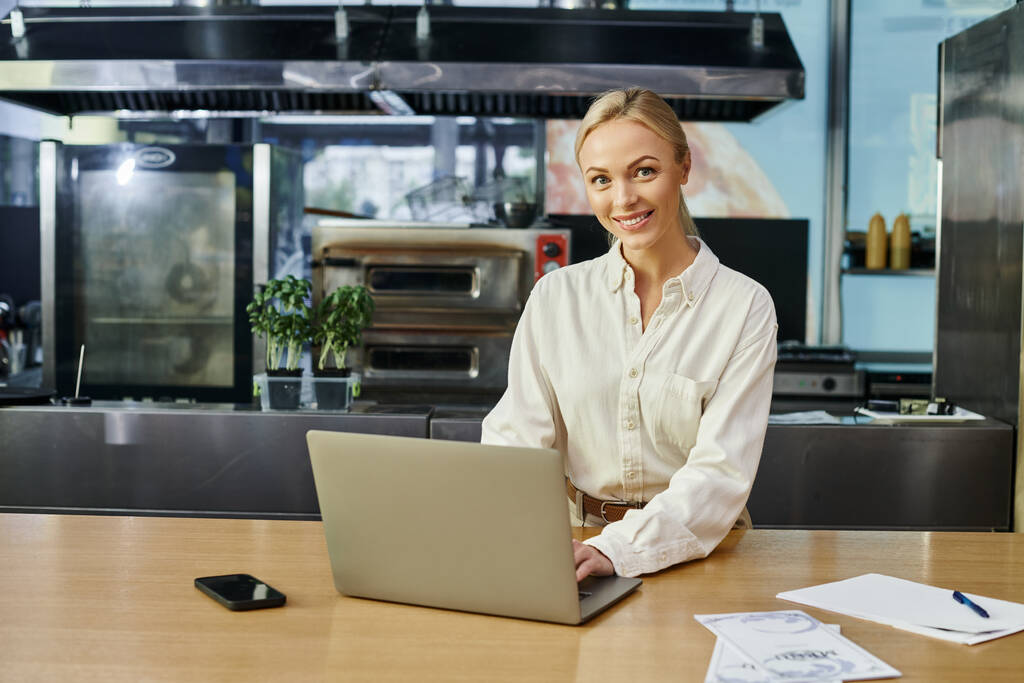 sonriente mujer rubia que trabaja en el ordenador portátil cerca de teléfono inteligente en el mostrador en la cafetería moderna, pequeña empresa - Foto, Imagen