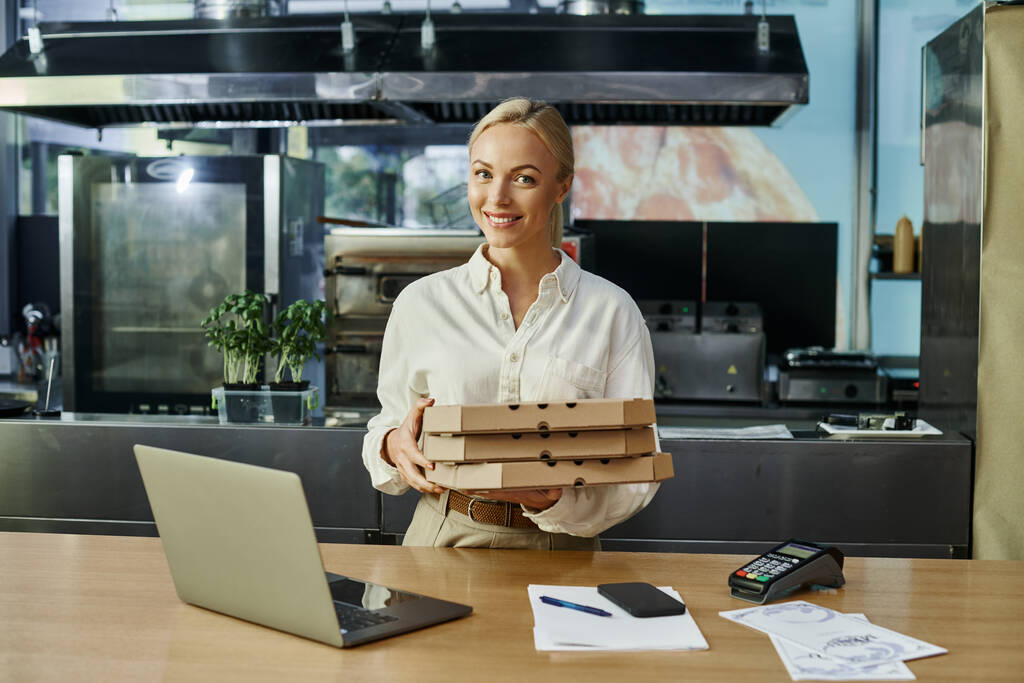 Lächelnde blonde Frau mit Pizzakartons in der Nähe digitaler Geräte und Zahlungsterminal am Tresen im Café - Foto, Bild