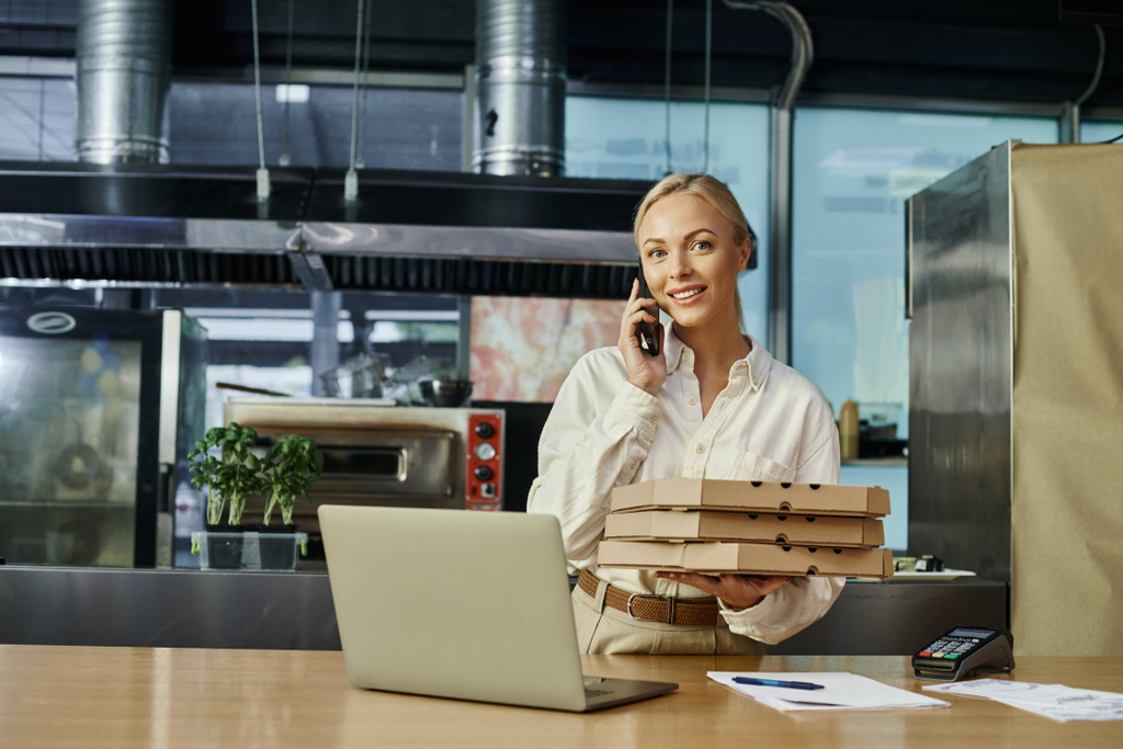 heureux jolie gestionnaire avec des boîtes à pizza parler sur smartphone près du terminal de paiement et ordinateur portable dans le café - Photo, image