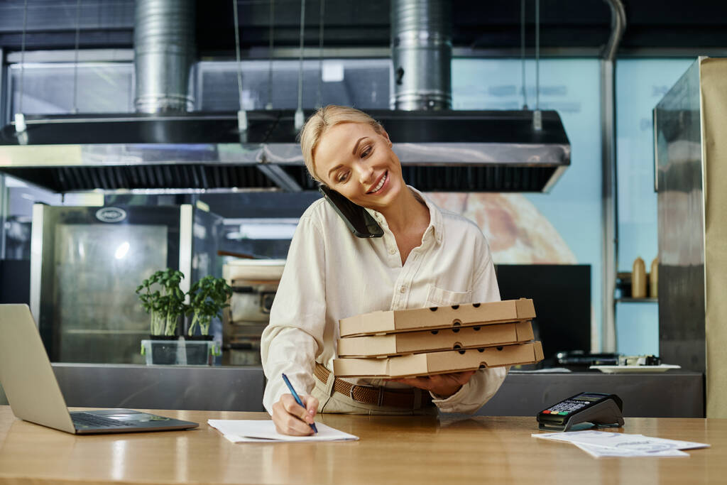 mujer rubia feliz con cajas de pizza hablando en el teléfono inteligente y el orden de escritura en el mostrador en la cafetería - Foto, imagen