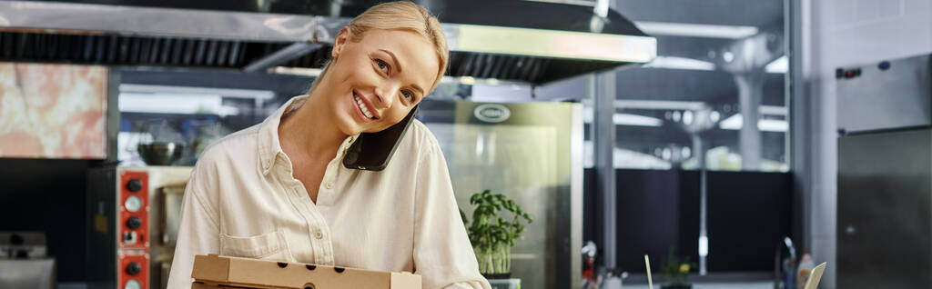 alegre gerente femenina con cajas de pizza hablando en el teléfono inteligente y en la cafetería moderna, pancarta horizontal - Foto, imagen