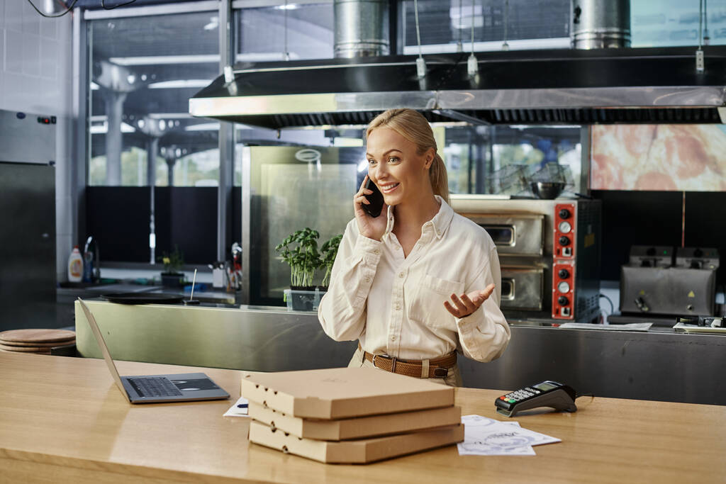 mujer rubia emocionada hablando en el teléfono inteligente cerca de cajas de pizza y portátil en el mostrador en la cafetería moderna - Foto, imagen