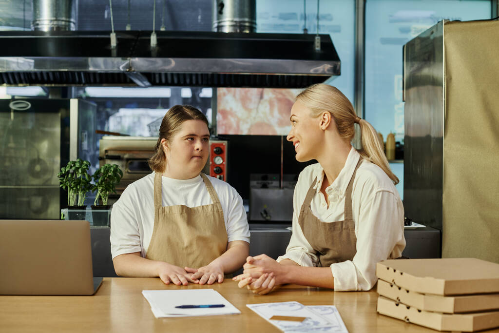 lachende cafe manager praten met jonge vrouwelijke werknemer met down syndroom in de buurt van laptop en pizza dozen - Foto, afbeelding