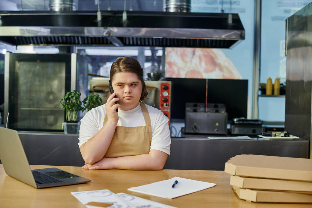 Nuori nainen alas oireyhtymä työskentelee kahvilassa ja puhuu älypuhelimella lähellä kannettavaa tietokonetta ja pizzalaatikoita - Valokuva, kuva