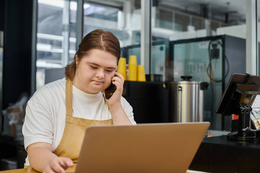 empleada femenina con síndrome de Down hablando en el teléfono inteligente cerca de la computadora portátil mientras trabaja en la cafetería moderna - Foto, Imagen