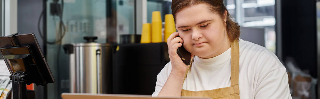 mujer joven con discapacidad mental hablando en el teléfono inteligente mientras trabaja en la cafetería moderna, pancarta - Foto, imagen