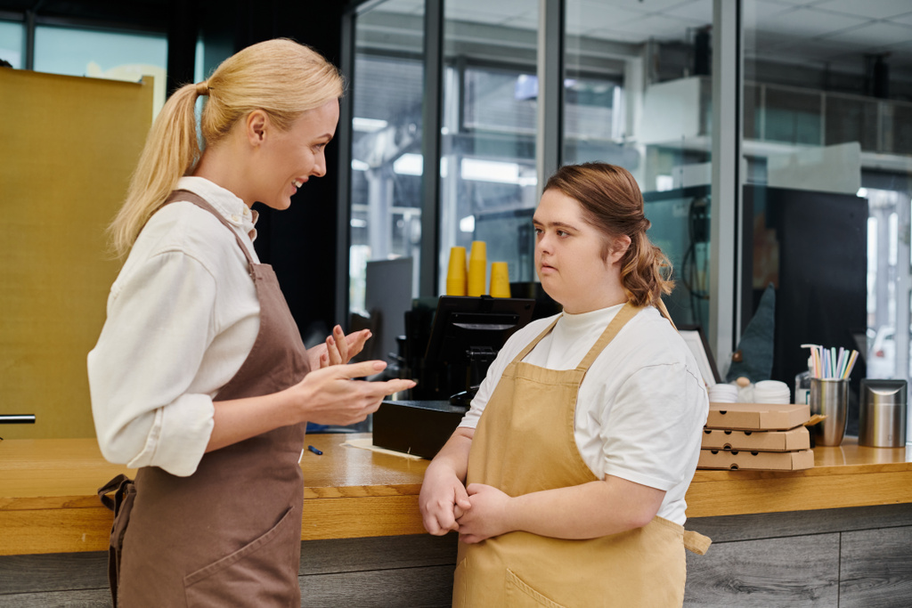 gerente sonriente hablando con una joven empleada con síndrome de Down en el mostrador en la cafetería moderna - Foto, Imagen