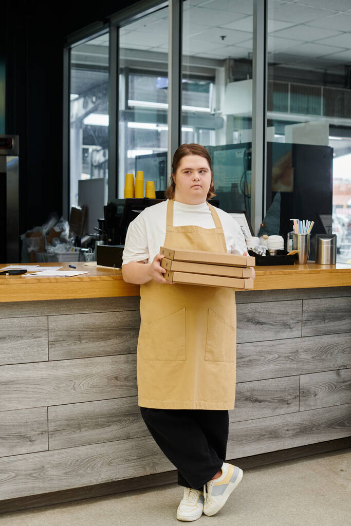 ダウン症の若い女性従業員は,現代のカフェのカウンターでピザ箱と立っています - 写真・画像