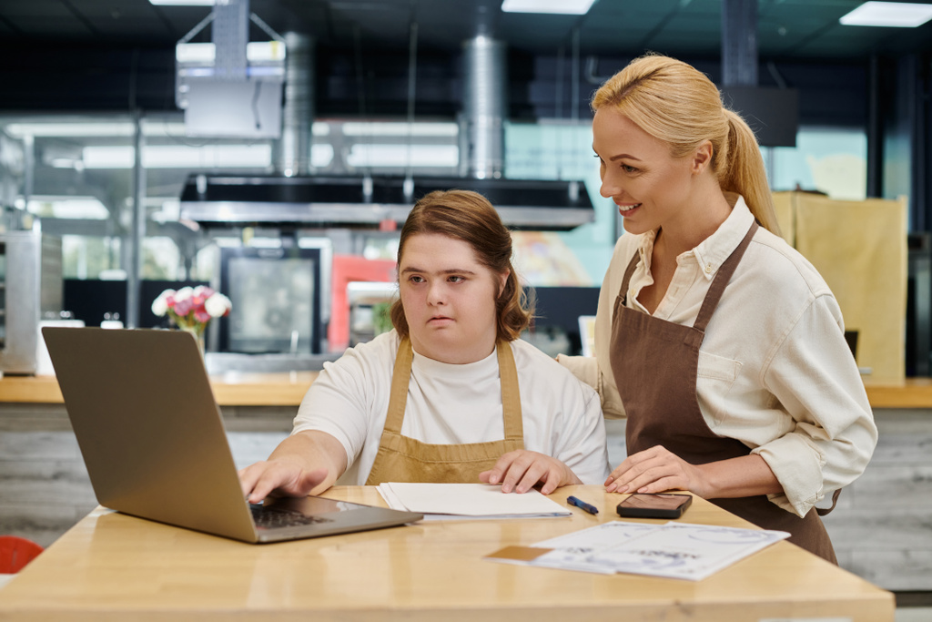 cheerful administrator smiling near female employee with down syndrome working in laptop in cafe - Photo, Image