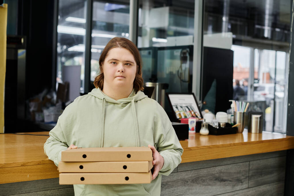 mladá žena s mentální poruchou držení krabice od pizzy a při pohledu na kameru v moderní útulné kavárně - Fotografie, Obrázek