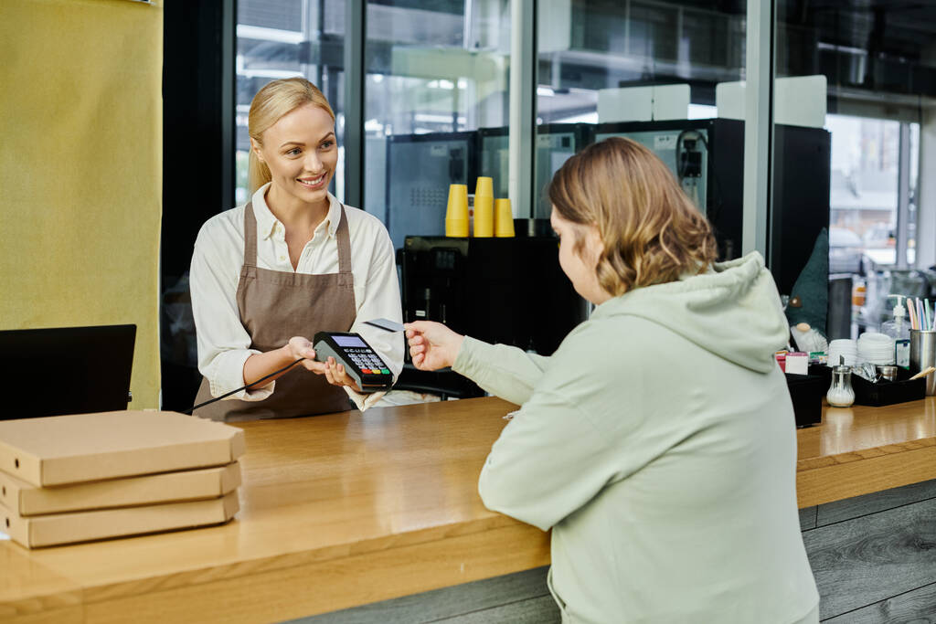 mujer joven con síndrome de Down pagar con tarjeta de crédito cerca de administrador con terminal en la cafetería - Foto, Imagen