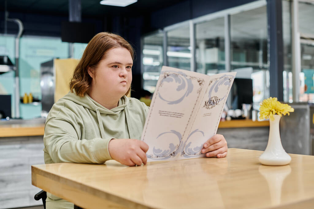 вдумчивая молодая женщина с синдромом Дауна смотрит на карту меню, сидя за столом в кафе - Фото, изображение