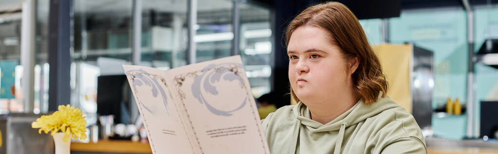 młoda kobieta z zespołem Downa trzymając kartę menu i myślenia w nowoczesnej kawiarni, baner poziomy - Zdjęcie, obraz