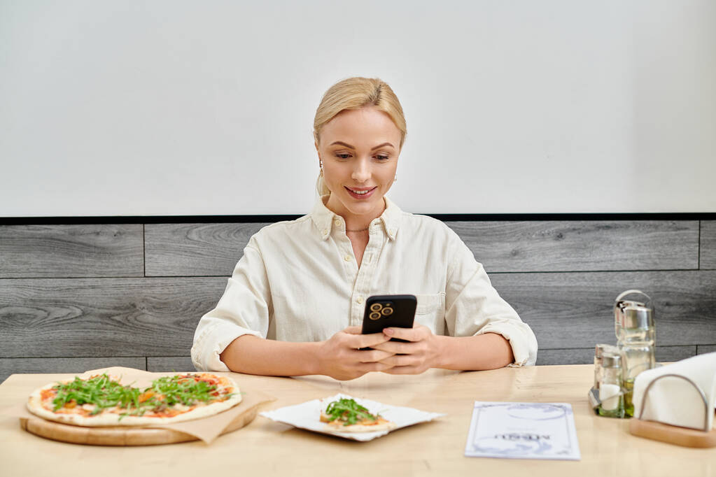 smiling blonde woman messaging on smartphone near delicious pizza on table in modern cozy cafe - Photo, Image