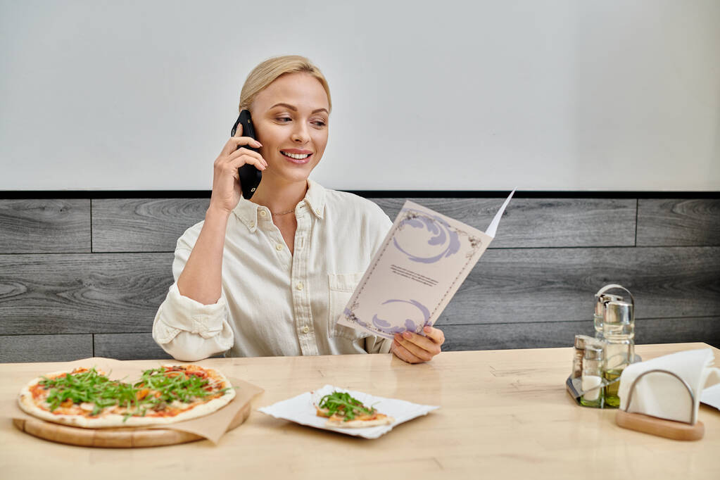 χαμογελαστή ξανθιά γυναίκα μιλάει στο smartphone και ψάχνει στο μενού κάρτα κοντά νόστιμο πίτσα στο καφέ - Φωτογραφία, εικόνα