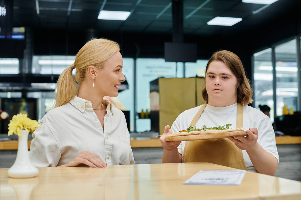 νεαρή σερβιτόρα με διανοητική αναπηρία κρατώντας νόστιμη πίτσα κοντά σε χαμογελαστή γυναίκα που κάθεται στο καφέ - Φωτογραφία, εικόνα