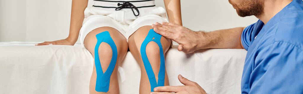 bebaarde arts die kinesiologische banden op de knie van zijn patiënt legt tijdens zijn afspraak, spandoek - Foto, afbeelding