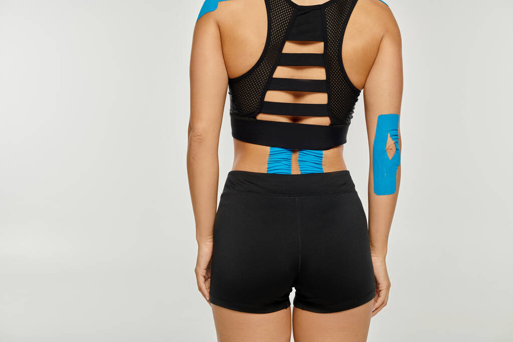 vue recadrée de la jeune femme en tenue de sport noire avec des bandes kinésiologiques sur le dos sur fond gris - Photo, image
