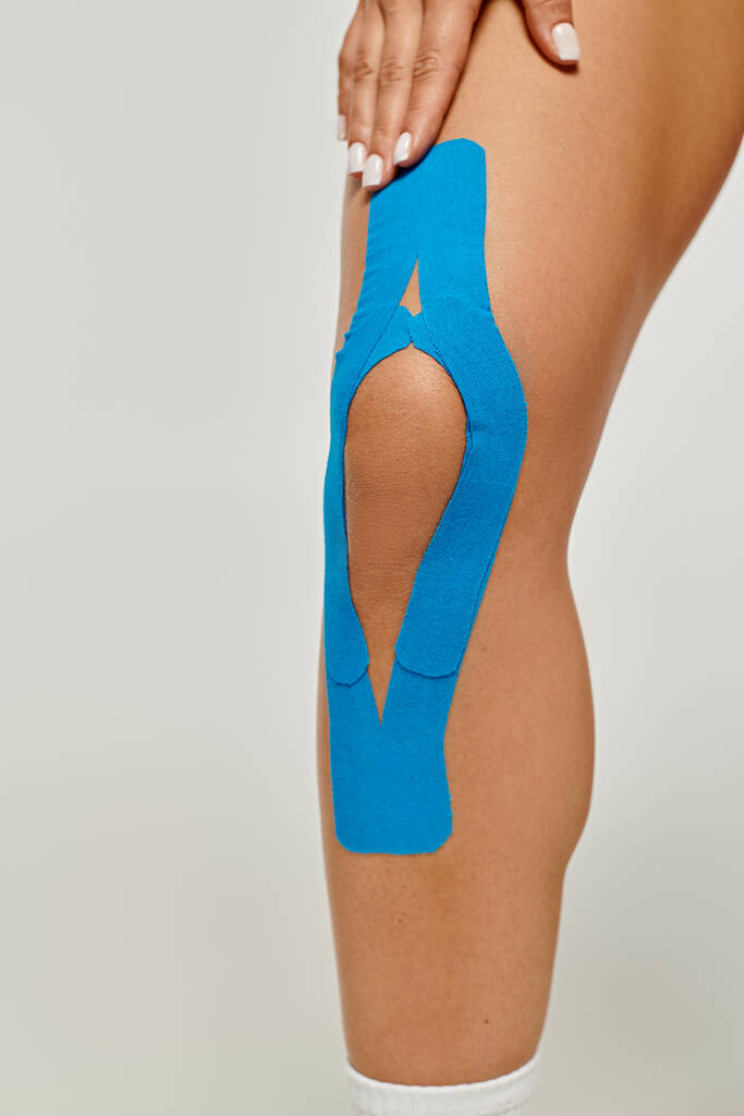 обрізаний вид молодої жінки з синіми кінезіологічними стрічками на її коліні позує на сірому фоні - Фото, зображення