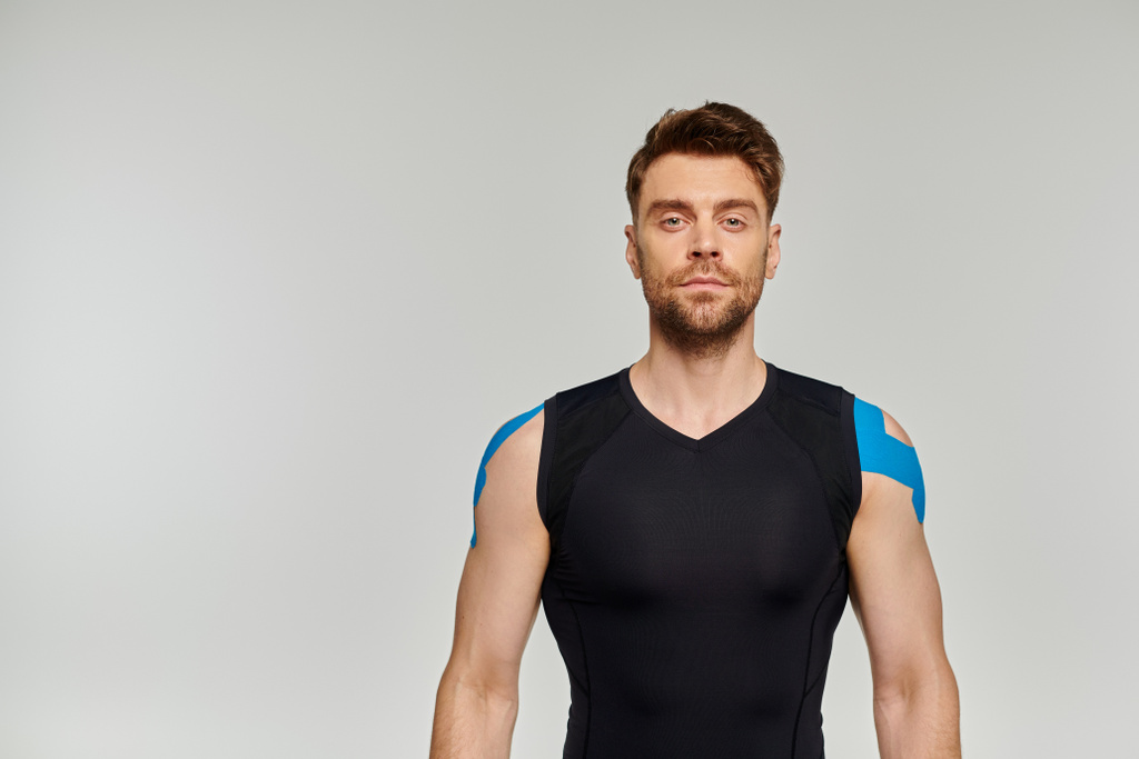 schöner bärtiger Mann in schwarzer Sportkleidung mit kinesiologischen Klebebändern am Körper, der in die Kamera blickt - Foto, Bild