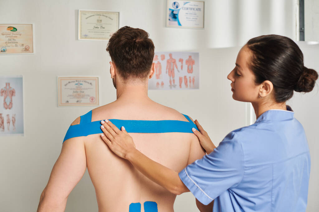 hermosa joven doctora poniendo cintas kinesiológicas en la espalda y hombros de su paciente, cuidado de la salud - Foto, imagen