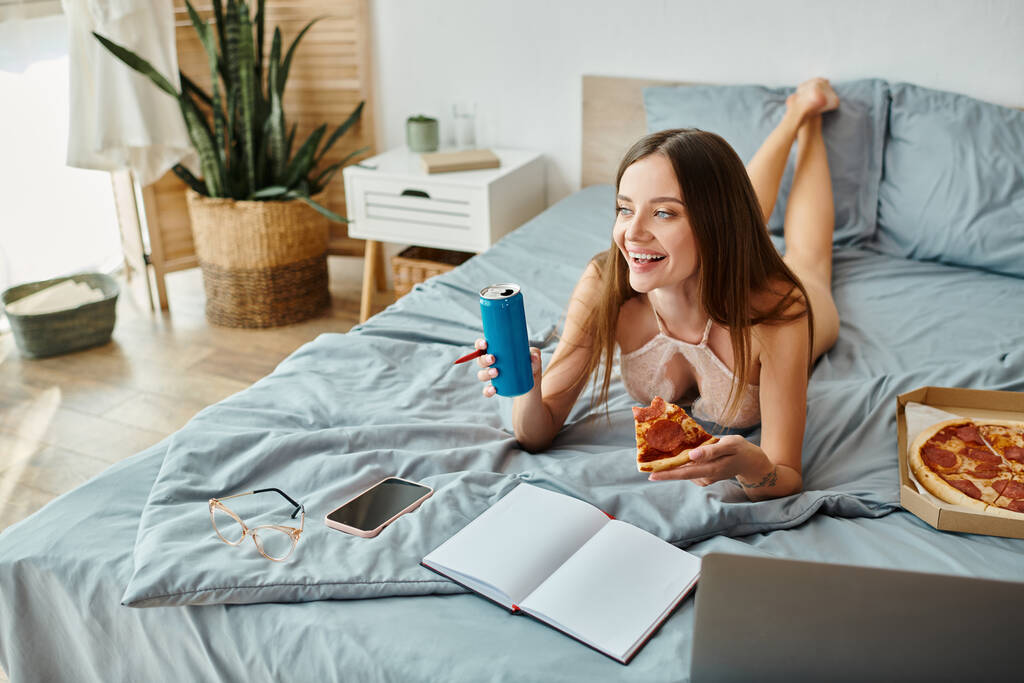 ansprechende lustige Frau, die mit Pizza und Limo im Bett liegt und wegschaut, während sie am Laptop arbeitet - Foto, Bild