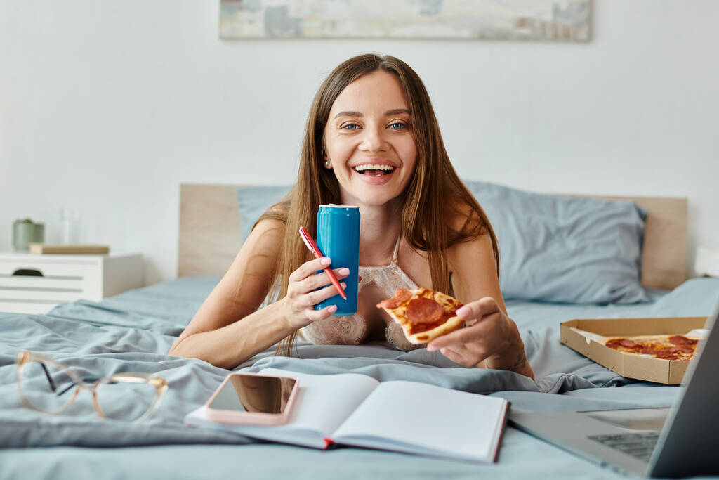 fröhliche, verführerische Frau mit langen Haaren, die mit Pizza und Limo im Bett liegt und in die Kamera blickt - Foto, Bild