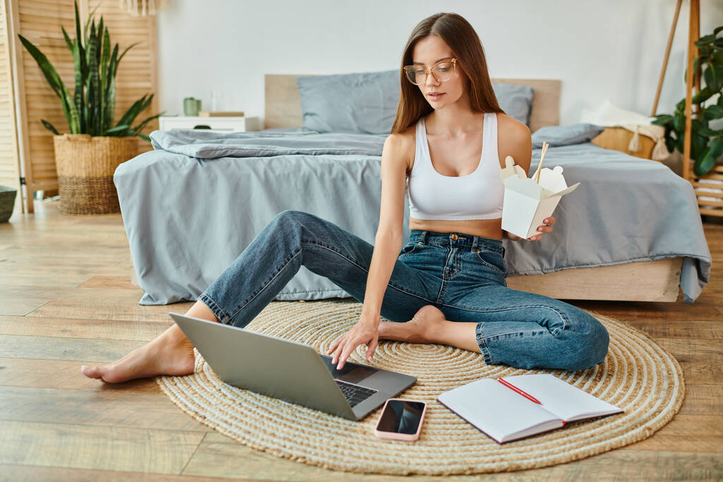 mujer atractiva pensativa en ropa de casa acogedora sentada en el suelo con fideos y trabajando duro en el ordenador portátil - Foto, Imagen