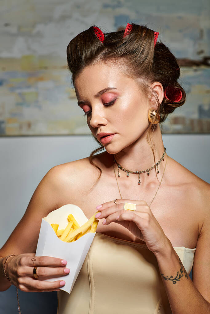 alluring young woman with hair curlers in beige corset looking at pack of fries in her hands - Photo, Image