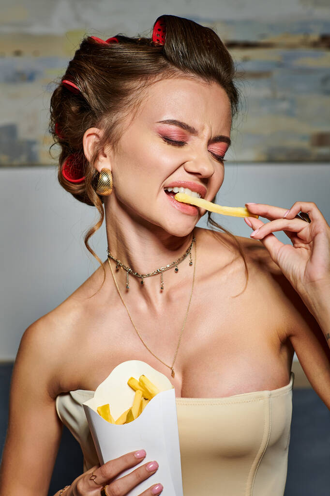 atractiva joven con rizadores de pelo en corsé beige sexy posando emocionalmente con papas fritas en las manos - Foto, imagen