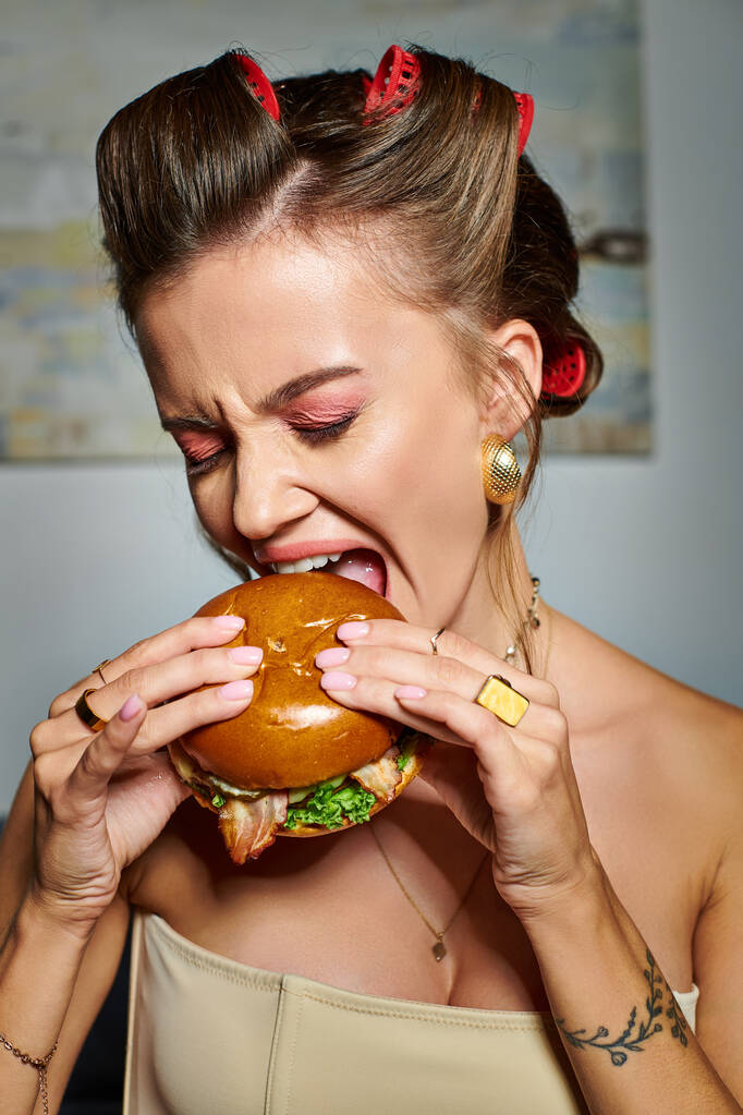 jonge mooie vrouw met krulspelden en accessoires eten heerlijke hamburger met eetlust - Foto, afbeelding