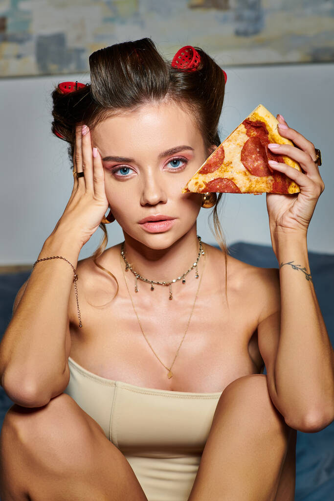 atractiva mujer sexy en seductor corsé beige posando con rebanada de pizza y mirando a la cámara - Foto, Imagen