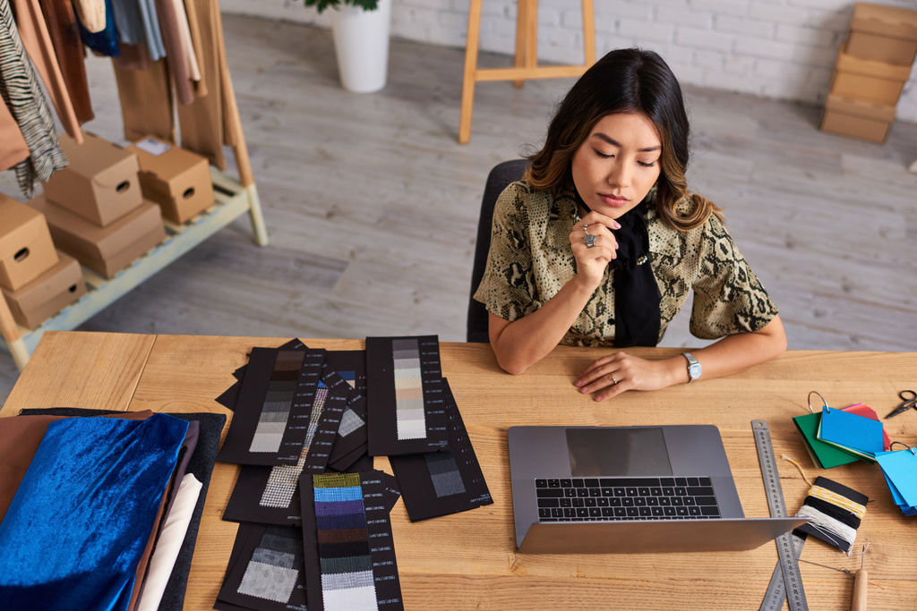 hoge hoek uitzicht van ernstige Aziatische mode ontwerper werken op laptop in de buurt van kleur monsters in atelier - Foto, afbeelding