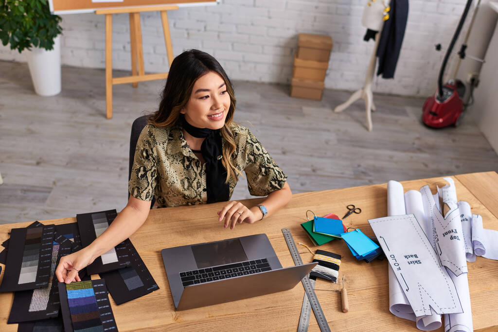 gelukkig aziatische mode stylist glimlachen in de buurt van laptop en kleur stalen met naaipatronen in atelier - Foto, afbeelding