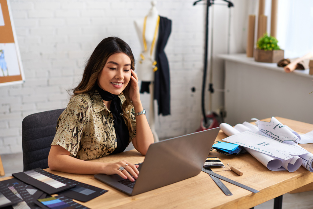 heureux asiatique créateur de mode travaillant sur ordinateur portable près de couture pattens et des échantillons dans son propre atelier - Photo, image