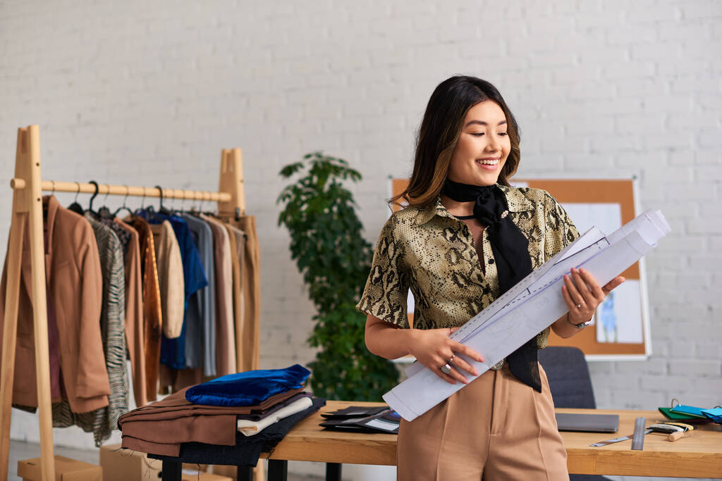 nuori iloinen aasialainen suunnittelija seisoo ompelu kuvioita työpöytä moderni henkilökohtainen atelier - Valokuva, kuva