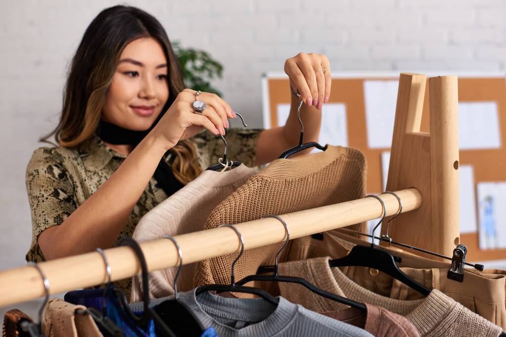spokojený úspěšný asijský stylista držící módní ležérní oděvy v osobním módním studiu - Fotografie, Obrázek
