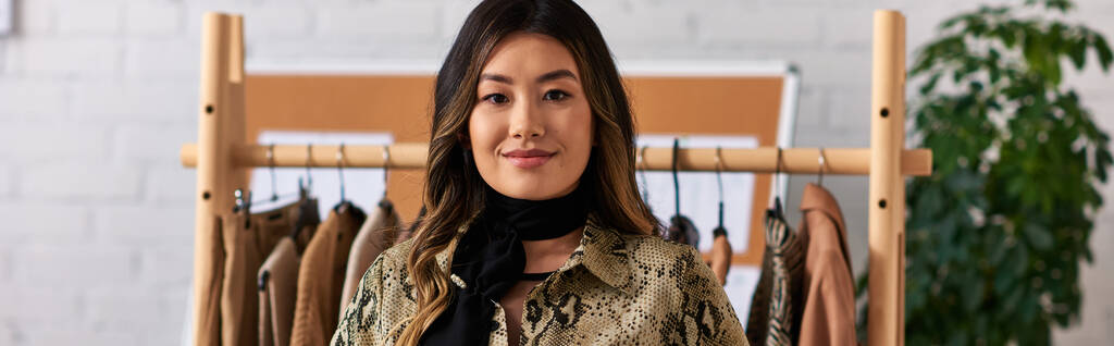 fröhliche asiatische persönliche Stylistin blickt in die Kamera in der Nähe von Regal mit maßgeschneiderten Kleidungsstücken im Atelier, Banner - Foto, Bild