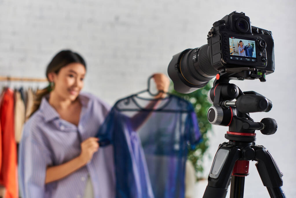 Сосредоточьтесь на цифровой камере рядом с молодым азиатским стилистом с модной блузкой в ателье, модный блог - Фото, изображение