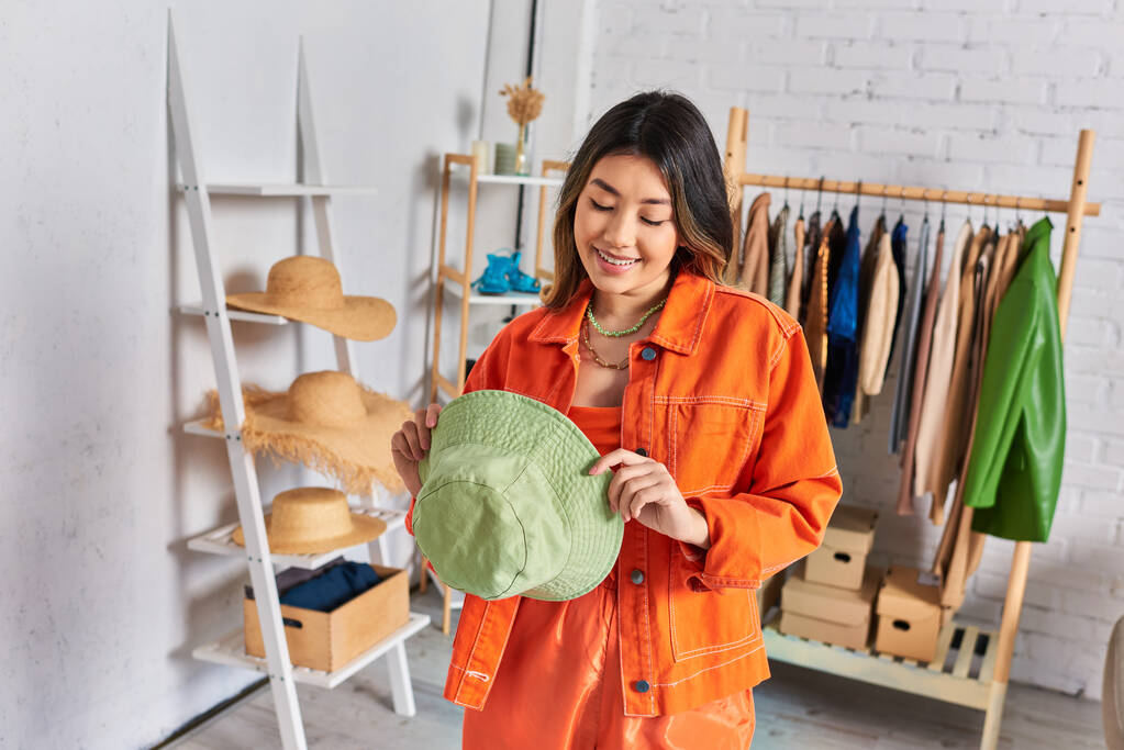 zufriedene asiatische Schneiderin in orangefarbener Kleidung mit Panamahut in privatem Atelier, Kleinunternehmen - Foto, Bild