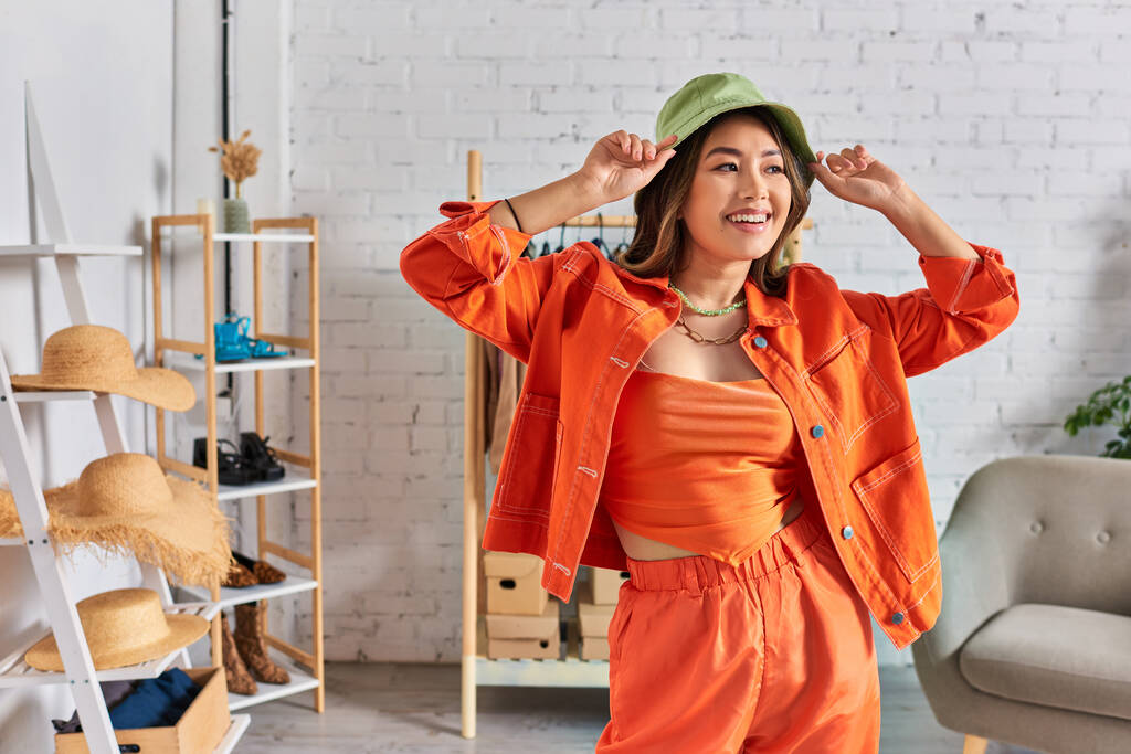 ενθουσιασμένοι ασιάτης σχεδιαστής σε πορτοκαλί ρούχα φορώντας panama καπέλο και κοιτάζοντας μακριά στο δικό του ατελιέ - Φωτογραφία, εικόνα