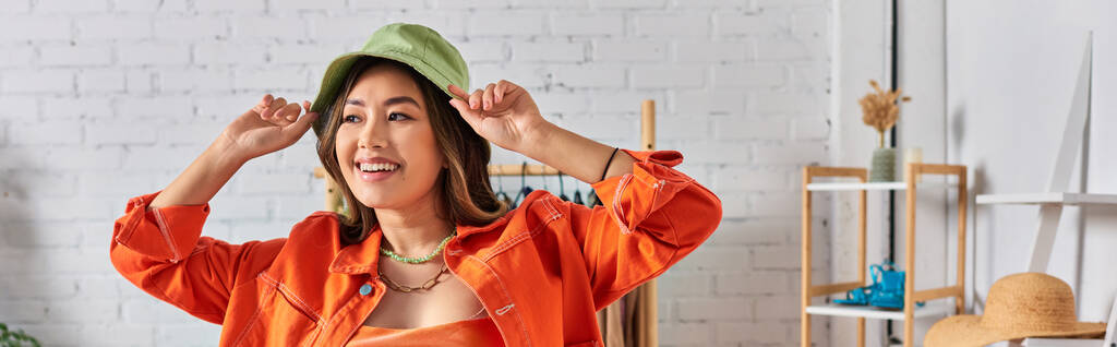 šťastný asijský stylista v oranžovém oblečení nosí panamský klobouk v oblečení ateliér, módní obchod, banner - Fotografie, Obrázek